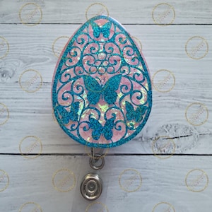 May include: A pink and teal butterfly-themed badge reel. The badge reel has a pink background with a teal filigree design and teal glitter butterflies. The reel has a silver metal clip and a clear plastic strap.
