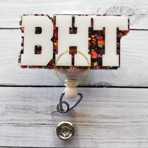 Peut inclure: Un enrouleur de badge avec un embout en acrylique blanc portant les lettres BHT dans une police grasse. Le fond de l'embout est rempli de paillettes épaisses orange, noires et argentées. Une sangle en plastique transparent avec un bouton-pression.