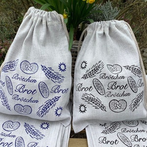Könnte beinhalten: Satz mit sechs weißen Leinen-Brottüten mit blauen und schwarzen Druckdesigns. Die Taschen zeigen die Worte "Brötchen Brot" und Bilder von Brot und Weizen.
