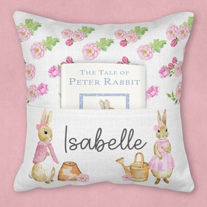 May include: A white pillow with a pocket for a book. The pillow has a floral pattern with pink roses and green leaves. The book is titled "The Tale of Peter Rabbit". The pillow also has two bunnies on it, one holding a watering can and the other holding a pot. The pillow is personalized with the name "Isabelle".