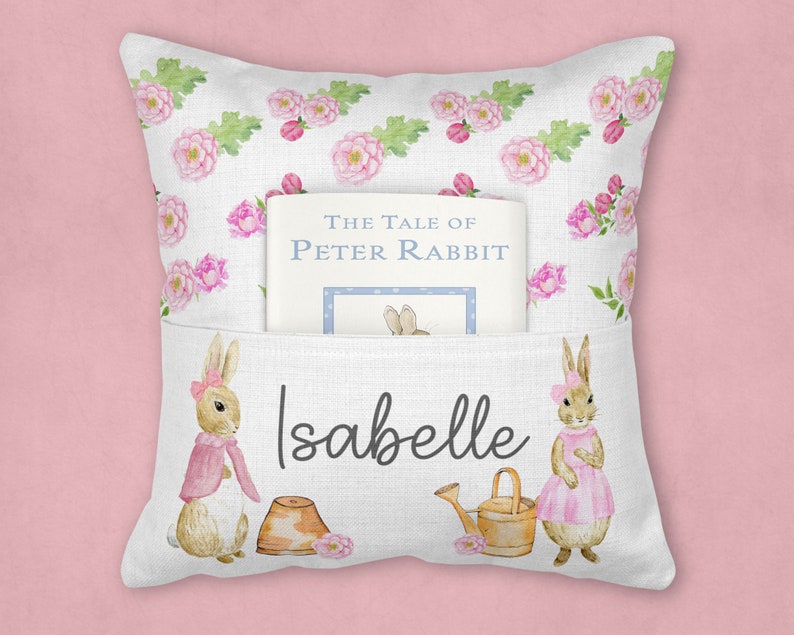 May include: A white pillow with a pocket for a book. The pillow has a floral pattern with pink roses and green leaves. The book is titled "The Tale of Peter Rabbit". The pillow also has two bunnies on it, one holding a watering can and the other holding a pot. The pillow is personalized with the name "Isabelle".