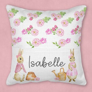May include: A white pillow with a pocket on the front. The pocket has a pink and green floral pattern with two bunnies on either side. The bunnies are wearing pink dresses and the text "Isabelle" is printed in black on the pocket.