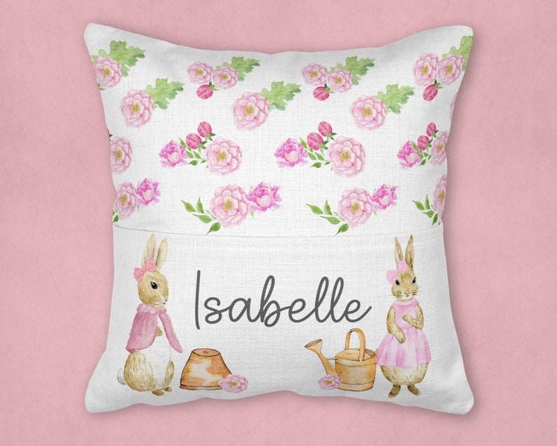 May include: A white pillow with a pocket on the front. The pocket has a pink and green floral pattern with two bunnies on either side. The bunnies are wearing pink dresses and the text "Isabelle" is printed in black on the pocket.