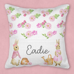 May include: A white pillow with a pocket features a floral pattern with pink flowers and green leaves. Two brown bunnies with pink accents are on either side of the pocket. The name "Eadie" is embroidered in black on the pocket.