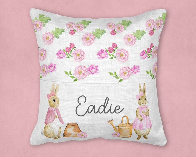 May include: A white pillow with a pocket features a floral pattern with pink flowers and green leaves. Two brown bunnies with pink accents are on either side of the pocket. The name "Eadie" is embroidered in black on the pocket.