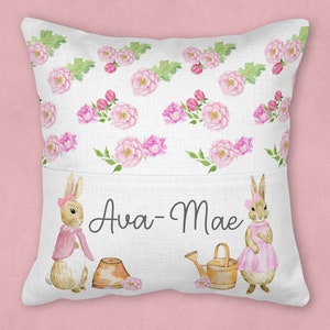 May include: A white pillow with a pocket on the front. The pocket has a pink floral pattern and two bunnies with pink accents. The text "Ava-Mae" is printed on the pocket.