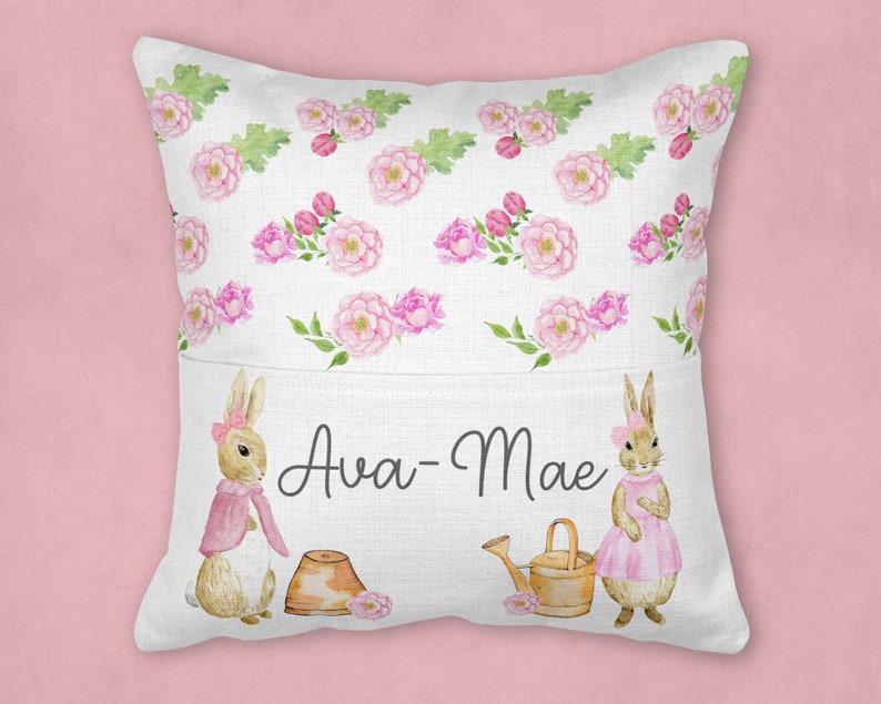 May include: A white pillow with a pocket on the front. The pocket has a pink floral pattern and two bunnies with pink accents. The text "Ava-Mae" is printed on the pocket.