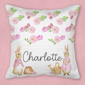 May include: A white pillow with a pocket features a floral pattern with pink flowers and green leaves. Two brown rabbits with pink accents are on the pocket, one holding a watering can and the other holding a pot. The text "Charlotte" is printed on the pocket.