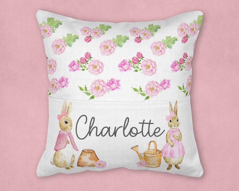 May include: A white pillow with a pocket features a floral pattern with pink flowers and green leaves. Two brown rabbits with pink accents are on the pocket, one holding a watering can and the other holding a pot. The text "Charlotte" is printed on the pocket.