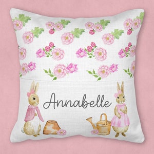 May include: A white pillow with a pocket on the front. The pocket has a pink and green floral pattern with two bunnies and the name Annabelle printed on it.