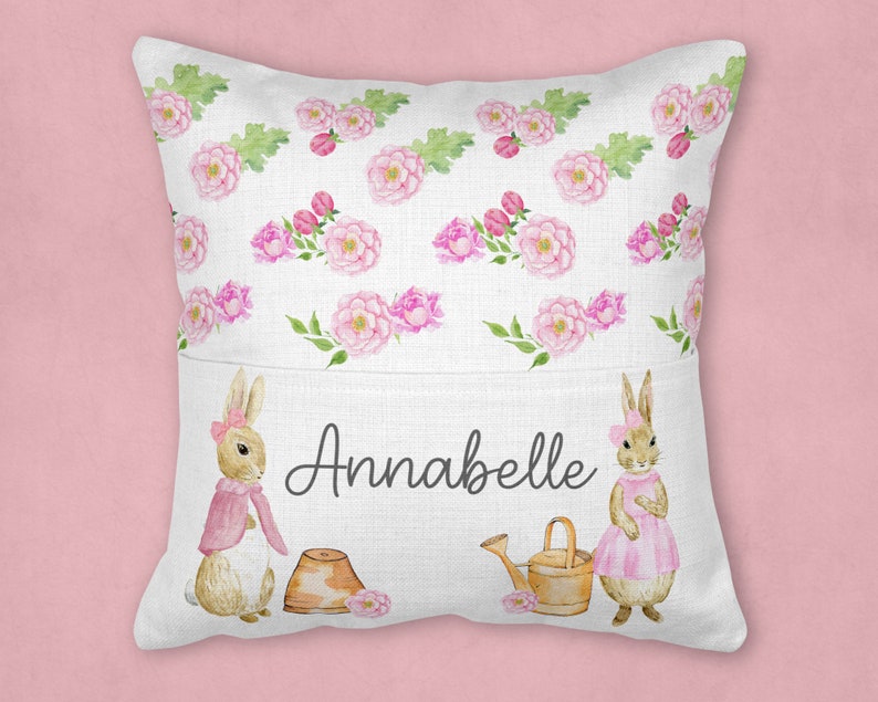 May include: A white pillow with a pocket on the front. The pocket has a pink and green floral pattern with two bunnies and the name Annabelle printed on it.