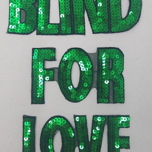 Puede incluir: Letras de lentejuelas verdes que deletrean "BLIND FOR LOVE" sobre un fondo blanco.