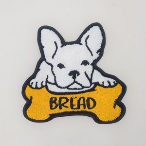 Può includere: Toppa ricamata bianca di un bulldog francese con la parola "BREAD" su un osso giallo.