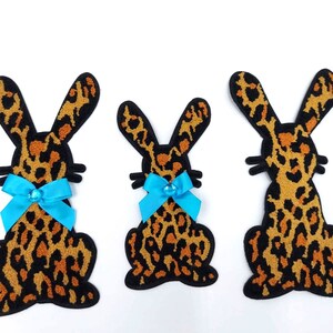 May include: Three plush leopard print bunny rabbit shapes with blue bows. The bunnies are different sizes and are facing forward.