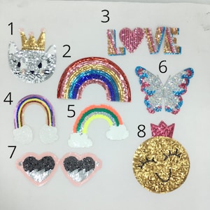 Puede incluir: Surtido de parches para planchar con una variedad de diseños, que incluyen un gato, arcoíris, una mariposa, un corazón y una cara sonriente. Los parches están decorados con lentejuelas de varios colores, incluidos rosa, azul, verde, amarillo y plateado.