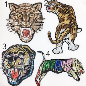 May include: Four embroidered iron-on patches featuring tiger designs. The patches are made of sequins and feature different colors and styles. The first patch is a tiger head with gold and brown sequins. The second patch is a tiger standing on its hind legs with gold and brown sequins. The third patch is a tiger head with black, gold, and purple sequins. The fourth patch is a tiger jumping with green, blue, pink, and gold sequins.