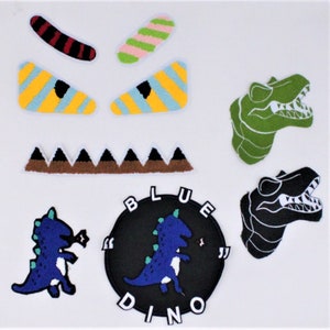 May include: A collection of colorful, embroidered patches featuring dinosaur and monster-themed designs. Includes a circular patch with the text "BLUE DINO", dinosaur heads, and various eye and mouth shapes. The patches are in shades of blue, green, black, yellow, and pink.
