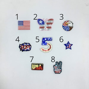 May include: Eight iron-on patches with patriotic themes. The patches feature a US flag, a butterfly, an eagle, a star, the word "USA", a peace sign, the number "5", and the word "HOORAY!" in a speech bubble. All patches are decorated with red, white, and blue colors.