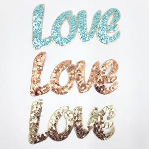 Könnte beinhalten: Drei Bügelbilder mit dem Wort "Love" in kursiv geschriebenen Buchstaben. Die Patches bestehen aus Pailletten in drei verschiedenen Farben: hellblau, gold und roségold.