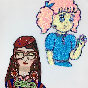Op de afbeelding: Twee geborduurde strijkpatches met cartoonmeisjes. Eén meisje heeft bruin haar, draagt een bril en een rode hoed met witte sterren. Het andere meisje heeft roze haar en een blauwe jurk.