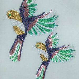 May include: Two sequined bird appliques in shades of gold, green, pink, and purple. The birds are in a flying pose with their wings spread out.