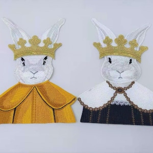 Könnte beinhalten: Zwei weiße gestickte Hasen-Patches mit goldenen Kronen und verschiedenen farbigen Roben. Der Hase links trägt eine gelbe Robe und der Hase rechts trägt eine schwarz-weiße Robe.