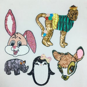 Könnte beinhalten: Fünf bunte Bügelbilder mit Tiermotiven. Die Patches sind aus Pailletten gefertigt und beinhalten einen rosa Hasen, einen goldenen Affen, ein braunes Reh, einen schwarzen Pinguin und ein graues Nashorn.