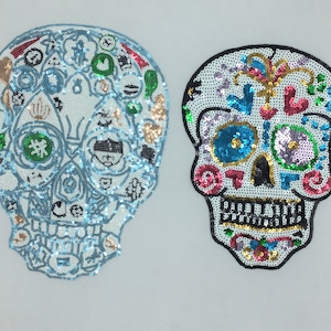 Puede incluir: Dos parches coloridos en forma de calavera con adornos de lentejuelas. Uno es azul y blanco con una variedad de pequeños diseños, el otro es blanco con contornos negros y lentejuelas de colores.