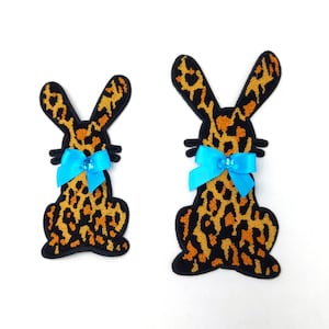 Puede incluir: Dos parches de conejo de fieltro con diseño de estampado de leopardo. Los conejos tienen contornos negros y lazos azules con un centro de pedrería.