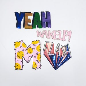 May include: A set of five colorful iron-on patches with various designs and text. The patches include the words "YEAH", "WAKE UP!" and "FLASH" in pink, white, and blue sequins. There is also a pink and yellow fuzzy letter "M" and a pink and blue diamond-shaped patch with the word "FLASH" in pink sequins.