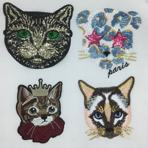 May include: Four embroidered iron-on patches featuring different cat designs. The first patch is a black and gold cat with green eyes. The second patch is a blue and white cat with pink star-shaped eyes and the word "paris" below. The third patch is a brown and gold cat with a crown and red roses. The fourth patch is a brown and white cat with blue eyes.