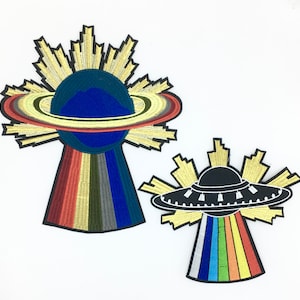 Könnte beinhalten: Zwei gestickte Bügelbilder. Eines zeigt einen Planeten mit Ringen und einem regenbogenfarbenen Schweif. Das andere zeigt eine schwarze fliegende Untertasse mit einem regenbogenfarbenen Schweif.