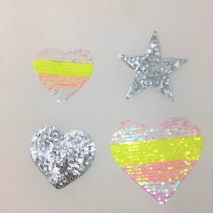 Puede incluir: Cuatro parches para planchar en forma de corazones y una estrella. Dos de los corazones son de color rosa y amarillo iridiscente con un contorno plateado. Los otros dos corazones y la estrella son plateados.