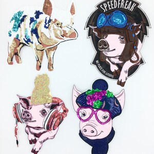 May include: Four embroidered iron-on patches featuring pig designs. The first patch is a white pig with blue and gold sequins. The second patch is a pink pig wearing a brown leather aviator helmet with blue goggles and the text "Speedfreak" and "No Place For Losers". The third patch is a pink pig wearing a gold sequined hat and headphones. The fourth patch is a pink pig wearing a blue beret with pink flowers and pink glasses.