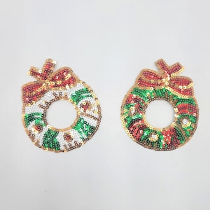 May include: Two sequined Christmas wreath ornaments. Each wreath is circular with a bow at the top. The wreaths are decorated with red, green, white, and gold sequins. The background is plain white.