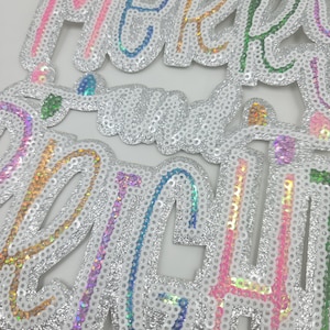 May include: A sequined decoration with the words "Merry Christmas" in a cursive font. The letters are outlined in various colors, including pink, blue, green, and gold, and are filled with iridescent sequins. The background is silver.