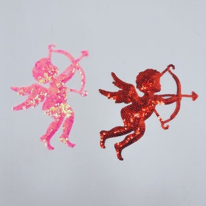 May include: Two sequined cupid figures, one pink and one red, holding bows and arrows. The figures are in a flying pose.