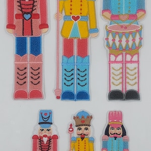 May include: Six embroidered nutcracker ornaments. The ornaments are all different designs, with some wearing hats, some holding drums, and some holding staffs. The ornaments are all in a variety of colors, including red, blue, yellow, pink, and white.