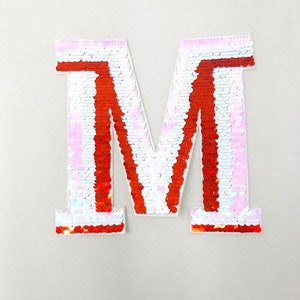 May include: A large, red and white sequined letter M. The letter is made of sequins that are arranged in a striped pattern. The sequins are iridescent and reflect light.
