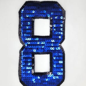 Op de afbeelding: Een blauwe nummer acht patch met pailletten en een zwarte rand. Het nummer heeft twee vierkante uitsparingen in het midden. De pailletten zijn felblauw en de patch staat op een witte achtergrond.