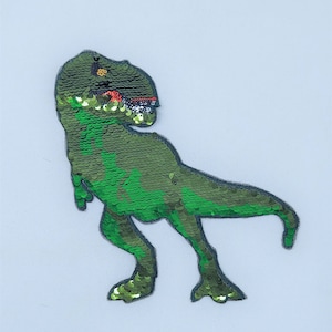Puede incluir: Parche de lentejuelas verde de un Tyrannosaurus Rex. El dinosaurio se representa en una pose caminando con la boca abierta, revelando detalles rojos y blancos. Las lentejuelas crean un efecto texturizado y brillante, y el parche está delineado en negro.