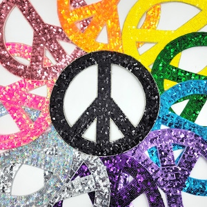 Op de afbeelding: Een verzameling kleurrijke peace-teken patches gemaakt van pailletten. De patches zijn in een cirkel gerangschikt met een zwart peace-teken in het midden.