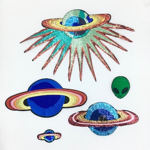 May include: Five colourful, sparkly, iron-on patches featuring planets and an alien. The patches are made of sequins and have a variety of colours, including blue, green, red, orange, and yellow.