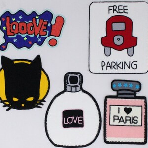 Puede incluir: Cinco parches para planchar con diferentes diseños. Un parche azul y rojo con la palabra "LOVE" en una burbuja. Un parche de silueta de gato negro. Un parche blanco con la palabra "LOVE" en un cuadrado rosa. Un parche rosa con las palabras "I LOVE PARIS" y un corazón. Un parche blanco con un coche rojo y las palabras "FREE PARKING".