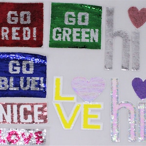 Puede incluir: Surtido de parches para planchar con varios colores, diseños y textos. Los parches incluyen "Go Red!", "Go Green", "Go Blue!", "Nice", "Love", "Hi", y corazones en rosa, morado e iridiscente.