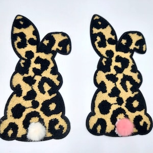Könnte beinhalten: Zwei plüschige Hasen-Patches mit Leopardenmuster. Ein Hase hat einen weißen flauschigen Schwanz und der andere einen rosa flauschigen Schwanz.