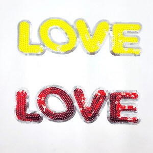 Könnte beinhalten: Zwei Bügelbilder mit dem Wort "LOVE" in großen, fetten Buchstaben. Das obere Patch ist gelb mit silbernen Pailletten, und das untere Patch ist rot mit silbernen Pailletten.