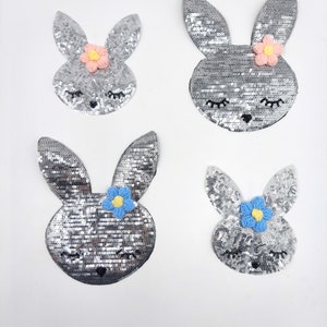Könnte beinhalten: Vier silberne Pailletten-Hasen-Patches mit gestickten Augen und einer Blume auf jedem. Die Blumen sind rosa, blau und hellblau.