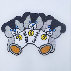 May include: Three grey and black teddy bears with blue eyes and fangs are sewn together. Each bear is wearing a white shirt with a yellow number six on it.
