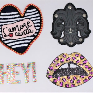 May include: Four iron-on patches with colorful designs. A blue and white striped heart with the text "'iamore canta" in pink. A black fleur-de-lis with silver studs. A multi-colored "HEY!" patch. A pink and black sequined lips patch with a leopard print border.